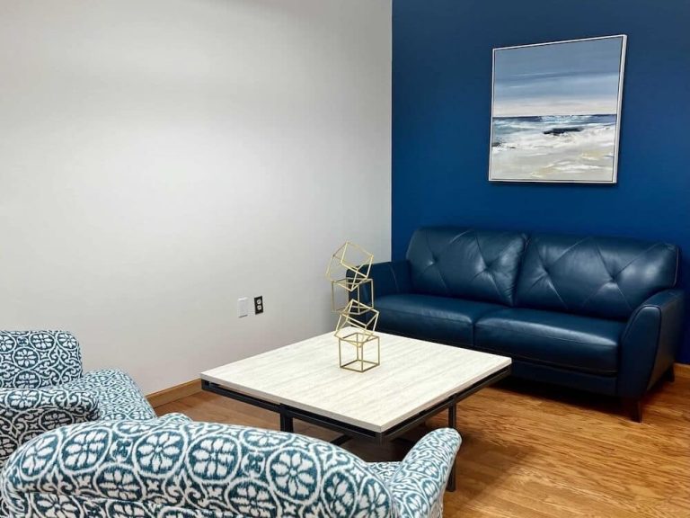 Modern therapy room with blue sofa, patterned chair, coffee table, and ocean artwork on a blue accent wall.