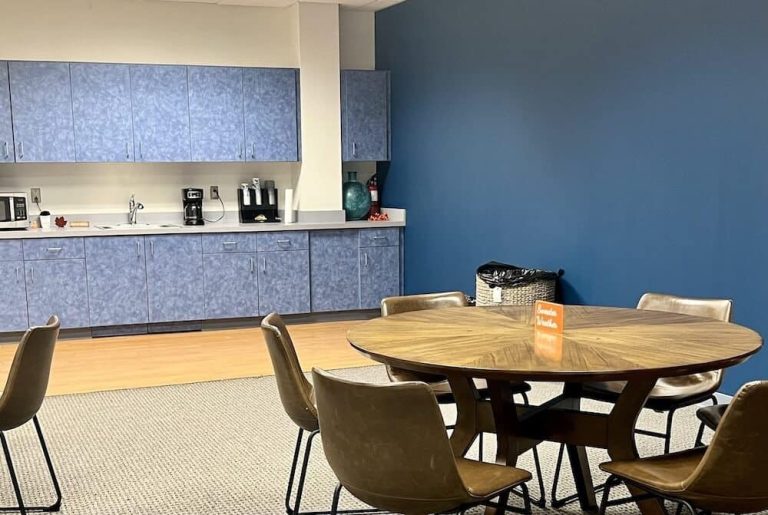 Break room with round table, chairs, and kitchenette with cabinets, sink, and coffee maker.