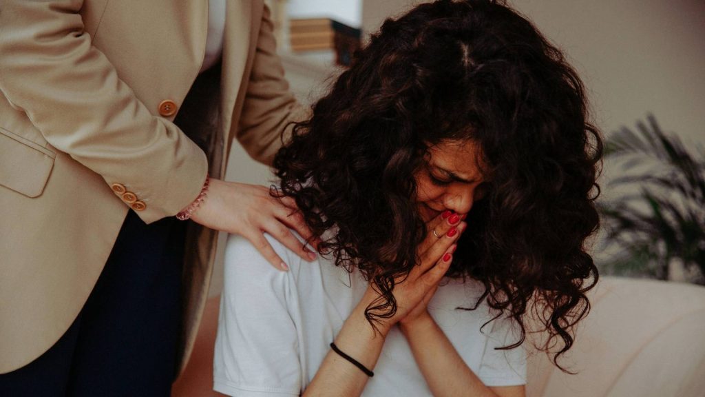 A person sitting with their head down while another person offers support.